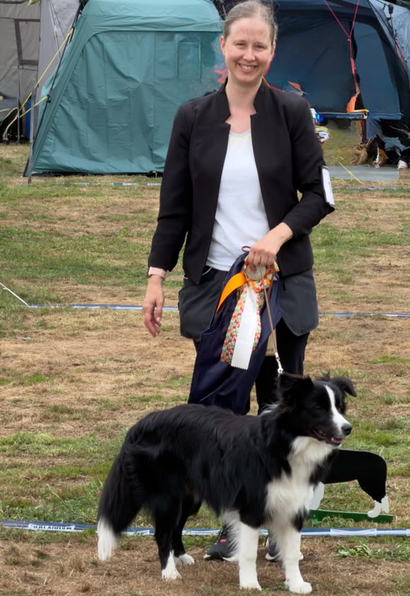 Eine Person steht auf einer Wiese neben einem schwarz-weißen Hund, im Hintergrund sind Zelte zu sehen.