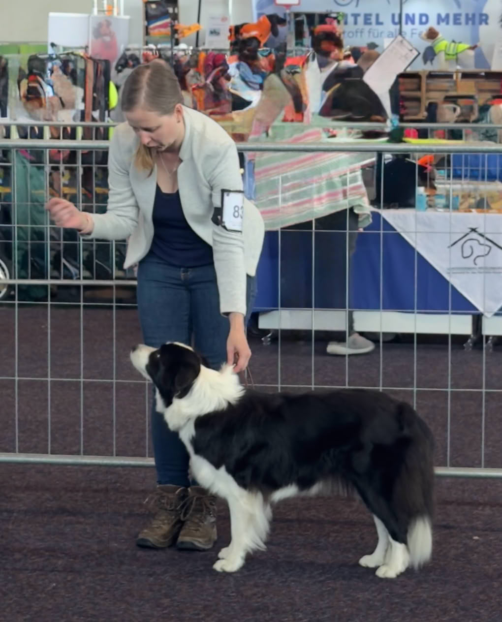 Eine Frau in einem hellen Blazer steht neben einem schwarz-weißen Hund in einer Ausstellungshalle.