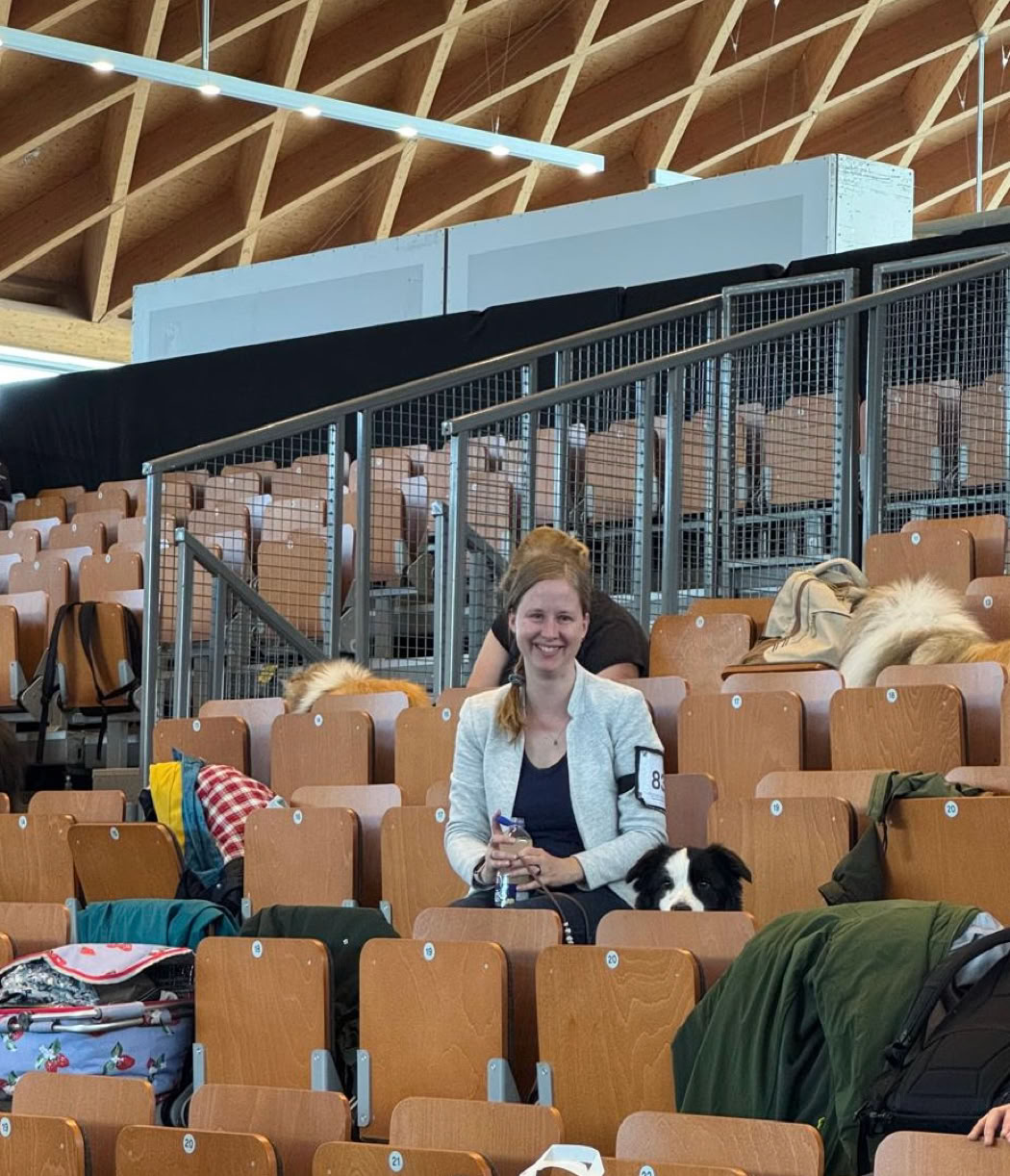 Eine Person sitzt in einem Hörsaal mit Holzbänken, umgeben von Taschen und Kleidung.